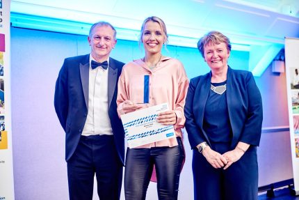 Berit Ritchie on stage receiving her award - with chair, Marie Burnham to her left and Chief Exec Mark Brooks to her right