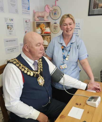 A man in ceremonial attire has his blood pressure checked by a nurse. They are seated at a table in a room with certificates on the wall.