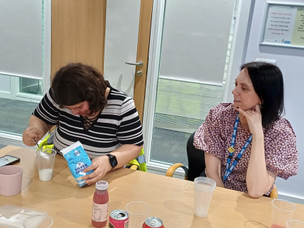 Person pouring spoon of sugar into a cup with another person watching