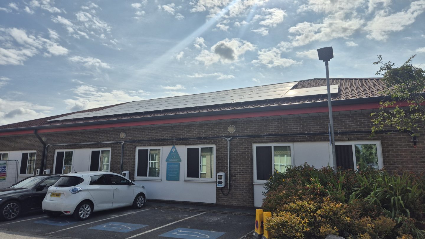 An image of a hospital ward from the outside, focus is on the roof with solar panels on. The sun is shining brightly on the panels
