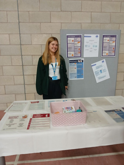 A woman stands beside a table displaying various posters, highlighting work at a physical health event.